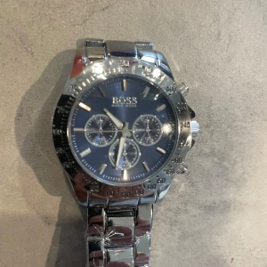 Silverfärgad klocka från BOSS - Stilren klocka från BOSS med blå urtavla och silverfärgat armband i metall. Klockan har tre mindre urtavlor, tydliga index och kronograf-funktion. Länken är robust och spännet är graverat med BOSS-loggan.