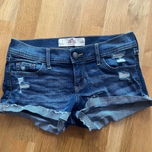 Blå jeansshorts från Hollister - Snygga blå jeansshorts från Hollister med slitna detaljer och fransig kant. Modellen är lågmidjad och har klassiska fickor både fram och bak med dekorativa sömmar. Perfekta för varma dagar!