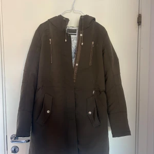 Olivgrön parkajacka med huva från Jofama - Säljer en olivgrön parkajacka från Jofama med huva och dragkedja framtill. Jackan har två bröstfickor med dragkedja och två fickor med lock och knapp. Fodret har ett grått blommigt mönster. Perfekt för kyligare dagar.Nypris: 1995 kr. Storlek 36-38.