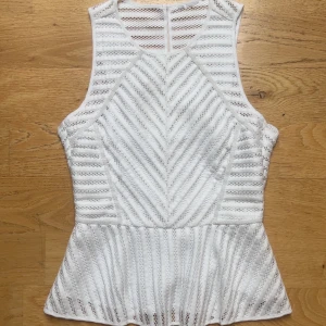 Vit topp - Säljer en vit ärmlös blus med spetsdetaljer och peplumskärning. Toppen har rund halsringning och dragkedja i ryggen. 