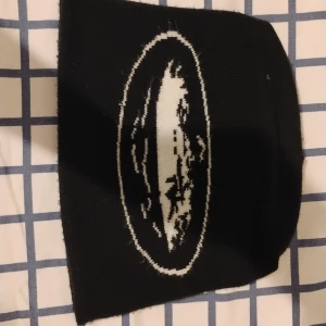 Svart stickad mössa med vitt ovalt mönster - Svart stickad mössa med ett vitt ovalt mönster framtill. Enkel och stilren design som passar till många olika outfits. Perfekt för kyligare dagar.