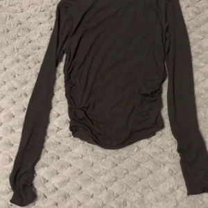 Mörkgrå långärmad topp från Vero Moda - En enkel mörkgrå långärmad topp från Vero Moda. Modellen är figurnära och har lätt rynkade detaljer i sidorna. Perfekt att matcha med jeans eller kjol för en stilren look.