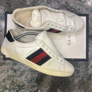 Vita sneakers från Gucci med röd och marinblå rand - Säljer ett par klassiska vita sneakers från Gucci med den ikoniska röd- och marinblå randen på sidan. Skorna har svart detalj vid hälen och är tillverkade i skinn. Perfekta för dig som gillar stilrena och lyxiga sneakers. Lägger till skosnören vid köp. Låda ingår 