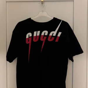 Svart t-shirt med Gucci-tryck - Cool svart t-shirt från Gucci med stort grafiskt tryck i vitt och rött på bröstet. Klassisk passform och rund halsringning. Perfekt för dig som vill sticka ut med en statement-tee.