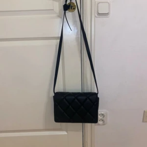 Svart quiltad axelväska - Säljer en stilren svart axelväska med quiltad design och lång axelrem. Väskan har magnetlås och flera fack inuti för enkel organisering. Perfekt för dig som vill ha en snygg accessoar till din outfit.