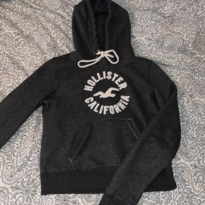 Mörkgrå hoodie från Hollister - Mörkgrå hoodie från Hollister med vit logga och texten 'Hollister California' på bröstet. Tröjan har huva med vita snören.