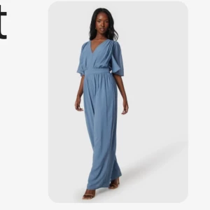 Blå lång jumpsuit med v-ringning från Vila - Säljer en snygg blå lång jumpsuit från Vila med v-ringad omlott-topp och vida ben. Jumpsuiten har korta ärmar och markerad midja för en smickrande siluett. Perfekt för dig som gillar stilrena och bekväma plagg.