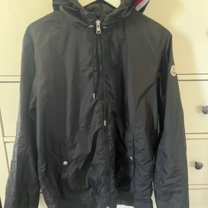 Moncler windbreaker  - Säljer en svart vindjacka från Moncler med huva och dragkedja framtill. Jackan har två framfickor med tryckknappar och Moncler-logga på ärmen. Perfekt för blåsiga dagar och enkel att matcha med det mesta.