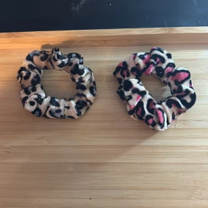 Två scrunchies med leopardmönster - Två scrunchies i glansigt tyg, båda med leopardmönster. Den ena är beige med svarta fläckar, den andra har inslag av rosa och svart på beige botten. Perfekta för att sätta upp håret med en trendig touch.