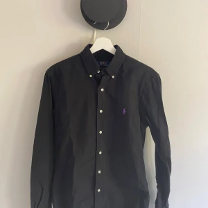 Svart skjorta från Polo Ralph Lauren - Klassisk svart skjorta från Polo Ralph Lauren med smal passform och knappstängning framtill. Liten broderad logga i lila på bröstet och krage med knappar. Perfekt för en stilren look.
