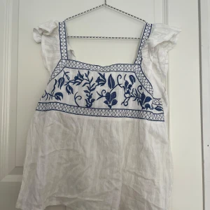 Vit blus  - Säljer en söt vit blus med blått broderat blommönster framtill och dekorativa axelband. Blusen har volangdetaljer vid axlarna och knytband i ryggen. Perfekt för en somrig look!🎀Funkar för dig med storlek s/m