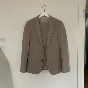 Beige kostym från Dressmann - Säljer en beige kostym från Dressmann i extra slim fit. Kavajen har två knappar framtill och klassiska slag. Byxorna är i matchande färg och har slim passform. Perfekt för student eller sommarens tillställningar.