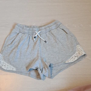 Grå shorts med spetsdetaljer från Lindex - Superbekväma grå shorts från Lindex med elastisk midja och vit dragsko. Snygga spetsdetaljer i vitt på sidorna ger en extra touch. Perfekta för varma dagar eller att chilla hemma i.