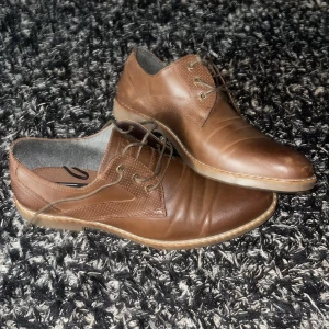 Bruna skinnloafers med snörning - Säljer ett par bruna loafers i skinn med snörning och rund tå. Skorna har en platt sula och diskreta sömmar som detalj. Perfekta för dig som gillar klassisk stil med en modern touch.