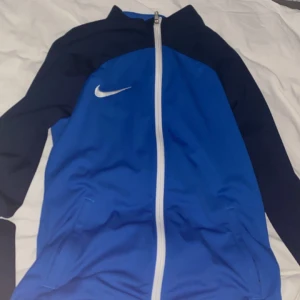 Blå och svart träningsjacka från Nike - Säljer en sportig blå och svart träningsjacka från Nike med vit dragkedja och logga på bröstet. Jackan har lång ärm och är perfekt för träning eller fotboll. Snygg och bekväm passform.