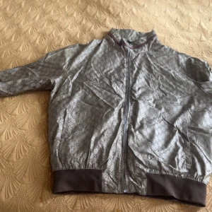 Gucci bomberjacka med monogram - Säljer en snygg bomberjacka från Gucci i beige med klassiskt GG-monogram över hela jackan. Jackan har bruna muddar, dragkedja framtill och hög krage med diskret röd detalj. Perfekt för dig som vill sticka ut med stil. Finns Luva och storlek 52