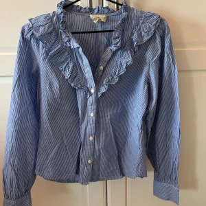 Blå- och vitrandig blus med volang från Holly & Whyte - Säljer en blå- och vitrandig blus från Holly & Whyte med volangdetaljer längs kragen och knäppning framtill. Blusen har långa ärmar och klassisk krage, perfekt för en stilren look. Nypris 400kr