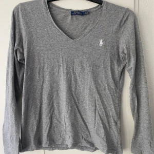 Grå långärmad tröja från Ralph Lauren - Säljer en grå långärmad tröja från Ralph Lauren med v-ringning och den klassiska vita loggan broderad på bröstet. Perfekt baströja med mjukt material och stilren design.