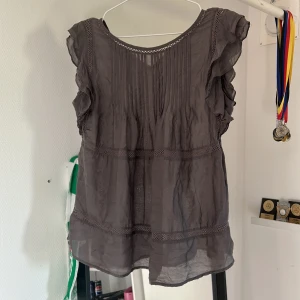 Mörkgrå blus med volangärmar - Gullig grå blus med fina volanger på axlarna. Blusen är gjort av ett fint och tunt tyg som ger en skön känsla mot huden. Super fin till sommaren! 