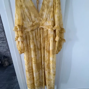 Gul långklänning med volang från NA-KD - Superfin gul långklänning från NA-KD med v-ringning och markerad midja. Klänningen har långa, lösa ärmar med dubbla volangdetaljer och knytning i ryggen. Tunt, flowigt material med subtilt mönster i olika gula nyanser.