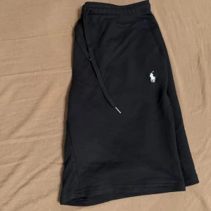 Grå och svarta shorts från Polo Ralph Lauren  - Snygga grå och svarta shorts från Polo Ralph Lauren med klassisk logga broderad på ena benet. De har snörning i midjan och är tillverkade i mjuk bomull, perfekta för avslappnade dagar.