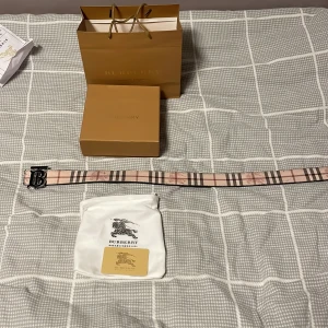 Beige och svart bälte från Burberry - Säljer ett klassiskt bälte från Burberry i beige med det ikoniska rutmönstret och silverfärgat spänne med Burberry-logga. Bältet är i skinn och levereras med originalpåse, ask och påse.