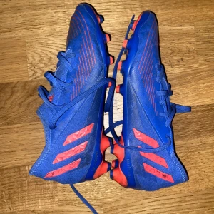 Blå och röda Adidas Predator fotbollsskor - Säljer ett par blåa Adidas Predator fotbollsskor med röda detaljer och snörning. Skorna har mönstrad ovansida och platt sula med dobbar för bra grepp på planen. Perfekta för dig som vill sticka ut på fotbollsplanen.