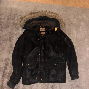 Svart vinterjacka med pälskant från Parajumpers - Säljer en svart vinterjacka från Parajumpers med huva och pälskant. Jackan har flera fickor, dragkedja och knappar samt detaljer som patch på ärmen och gul rem vid halsen. Perfekt för kalla dagar.