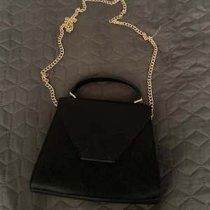 Svart handväska med guldkedja - Snygg svart handväska med stilren design och guldfärgad kedja som axelrem. Väskan har ett topphandtag och en elegant triangelformad frontflik. Perfekt accessoar för att lyfta din outfit.