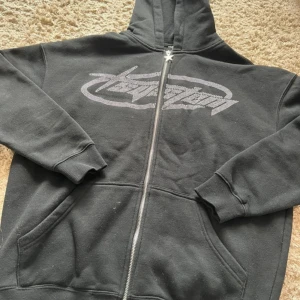 Svart hoodie med strasstryck från FSBN - Säljer en svart hoodie från FSBN i oversized modell med glittrigt strasstryck framtill. Dragkedja med stjärnformad zip och huva. Perfekt för dig som gillar streetstyle och vill ha något unikt!