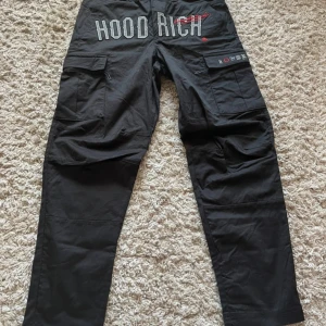 Svarta cargobyxor från Hoodrich - Svarta cargobyxor från Hoodrich med stora broderade loggor och textdetaljer på bakfickorna. Byxorna har flera fickor med lock och knappar samt snörning i midjan för extra passform. Perfekta för en streetwear-look.