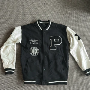 Svart och vit varsityjacka med broderade detaljer - Säljer en svart och vit varsityjacka med stora broderade bokstäver och siffror. Jackan har svart kropp, vita ärmar och broderad text på bröstet och ryggen. Ribbstickade muddar och knäppning framtill. Perfekt för en sportig look.