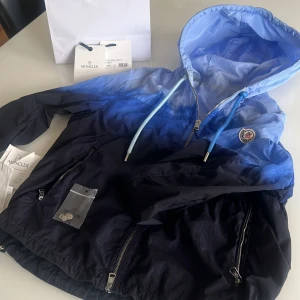 Blå vindjacka från Moncler - Säljer en snygg blå vindjacka från Moncler med huva och dragkedja. Jackan har en gradient från ljusblått upptill till mörkblått nedtill, två fickor med dragkedja och justerbara snören i huvan. Kom med ett pris denna Moncler säljs inte längre, Lämnar in den i Kemi tvätt innan den köps så den blir helt ren.