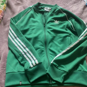Grön Adidas track jacket med vita ränder - Säljer en klassisk grön Adidas track jacket med vita ränder längs ärmarna och broderad logga på bröstet. Jackan har dragkedja framtill och ribbade muddar. Perfekt för en sportig och retro look.