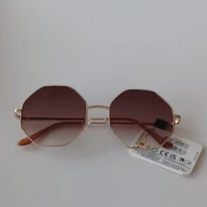 Guldiga åttakantiga solglasögon från Foster Grant, Prestige 7 - Snygga solglasögon med åttakantiga bruna glas och tunn guldfärgad metallbåge. Märket är Foster Grant och modellen heter prestige. Glasen har 100% UVA- och UVB-skydd. Perfekt accessoar för soliga dagar.