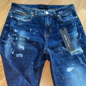 Helt nya jeans från Supply & Demand - Ett Aldrig andvändt par blå jeans från Supply & Demand med slitna detaljer och vit färgstänk. Klassisk femficksmodell med dragkedja och knapp. Raka ben och cool streetstil, perfekt för dig som gillar en trendig look.