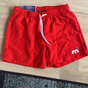 Röda badshorts från Mistral - Snygga röda badshorts från Mistral med vit snörning i midjan och liten logga på ena benet. Klassisk passform och elastisk midja, perfekta för stranden eller poolen.