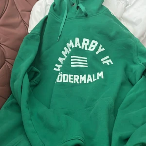 Grön hoodie från Hammarby IF - Grön hoodie med vit text 'HAMMARBY IF SÖDERMALM' och tre ränder på bröstet. Klassisk huva med dragsko och stor magficka framtill. Perfekt för dig som vill visa ditt stöd för Hammarby.