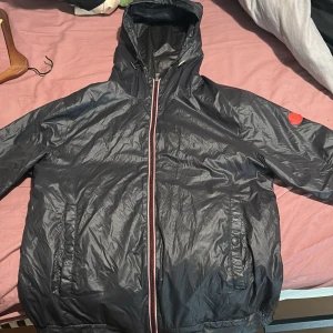 Svart vindjacka från Moncler med huva - Säljer en svart vindjacka från Moncler med huva och röd/vit dragkedja. Jackan har två fickor framtill och ett Moncler-märke på ärmen. Insidan har coolt Moncler-tryck och serietidningsdetalj. Perfekt för dig som vill ha en stilren och sportig look.