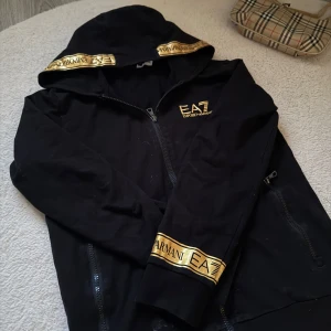 Svart hoodie från Emporio Armani EA7 med guldiga detaljer - Svart hoodie med dragkedja från Emporio Armani EA7. (Lite misssfärgad i luvan, men inget som syns på!)