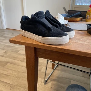 Arigato - Säljer ett par svarta slip-on sneakers med tjock vit platt sula. Skorna har en minimalistisk design utan snörning och är gjorda i mocka. Perfekta för en clean och stilren look.