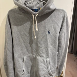 Grå hoodie från Polo Ralph Lauren - Klassisk grå hoodie från Polo Ralph Lauren med broderad logga på bröstet. Tröjan har huva med dragsko och en stor magficka framtill. Tillverkad i mjuk bomullsmix, perfekt för en avslappnad stil.