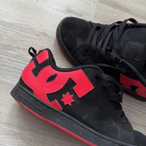 Svarta DC sneakers med rosa logga - Säljer ett par svarta DC sneakers med stor rosa DC-logga på sidan och rosa detalj på sulan. Skorna har snörning och platt sula, perfekt för dig som gillar streetwear och skate-stil.