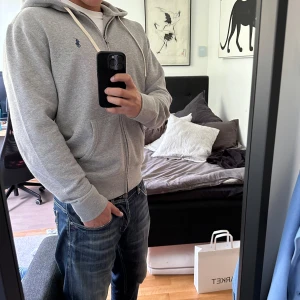 Grå hoodie med dragkedja från Polo Ralph Lauren - Säljer en klassisk grå hoodie från Polo Ralph Lauren med dragkedja och vit huva. Tröjan har långa ärmar, ribbade muddar och den ikoniska blå loggan broderad på bröstet. Perfekt för en avslappnad stil. Den är i storlek L men passar mig som vanligt viss har M