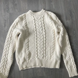 Vit stickad tröja med flätmönster - Säljer en vit stickad tröja med klassiskt flätmönster och rund hals. Tröjan har långa ärmar och ribbade muddar vid ärmslut och nederkant. 