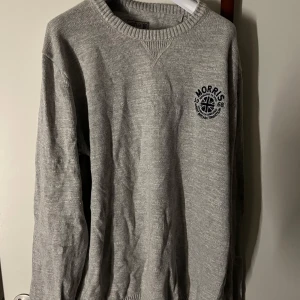 Morris tröja - Snygg grå stickad tröja från Morris med broderad logga på bröstet. Tröjan har rund halsringning och ribbade muddar vid ärmslut och nederkant. Perfekt för en stilren och avslappnad look.