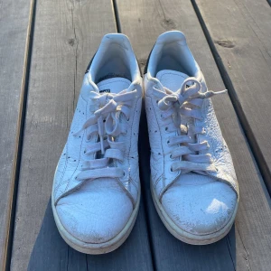 Vita Adidas Stan Smith sneakers - Klassiska vita Adidas Stan Smith sneakers med blå/svart detaljer på hälen och perforerade ränder på sidorna 💓. Skorna har snörning och är tillverkade i skinn med platt sula och rund tå. Perfekta för en stilren look och passar till allt 💕. De har flera tecken på slitage. Här har de ingen sula i men finns sedan som kommer med 🥰