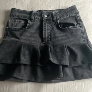 Svart jeanskjol med volang - Säljer en svart jeanskjol med volangdetalj nertill. Kjolen har klassiska jeansfickor fram och bak samt knapp och dragkedja i midjan. Perfekt för dig som gillar en edgy men söt stil.