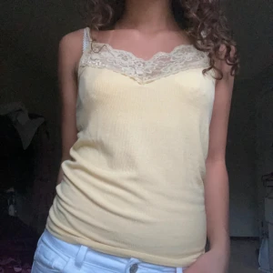 Gul ribbad linnetopp med spets - Säljer en ljusgul ribbad linnetopp med smala axelband och vacker spetsdetalj vid urringningen. Perfekt för varma dagar eller som ett lager under en skjorta. Superfin och enkel att styla till olika looks!