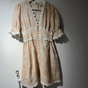 Mönstrad kortklänning från By Malina - Superfin kortklänning från By Malina i ljusa pastellfärger med paisleymönster. Klänningen har v-ringning, knappar framtill, spetsdetaljer och korta puffärmar. Perfekt för sommarens tillfällen! Väldigt förmånligt pris så först till kvarn!